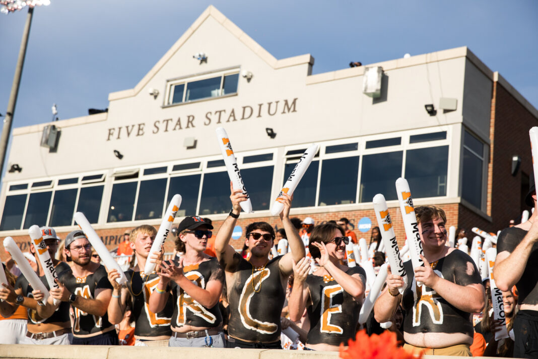 A group of fans with painted chests and foam sticks cheer in front of Five Star Stadium on a sunny day.