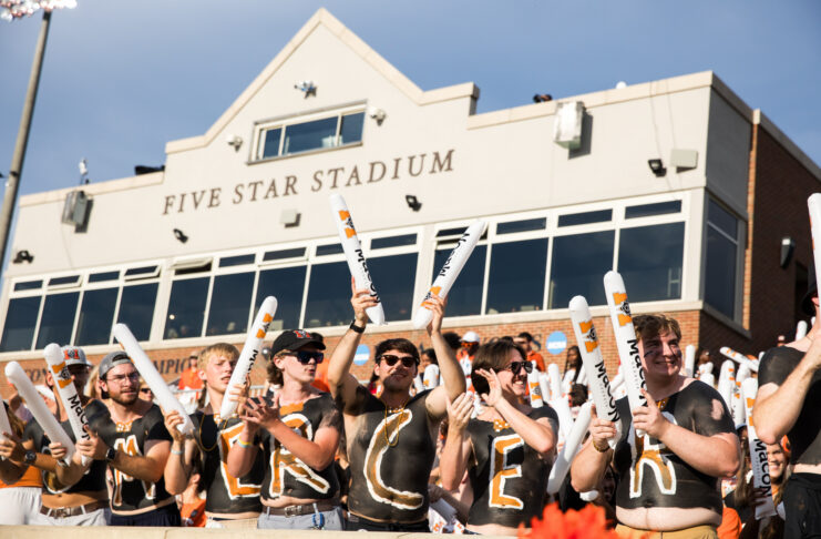 A group of fans with painted chests and foam sticks cheer in front of Five Star Stadium on a sunny day.