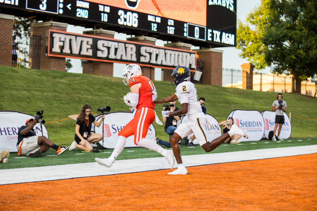 Football player in orange catches pass in end zone as defender in white trails behind at Five Star Stadium.