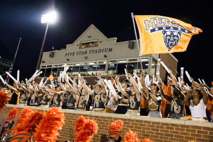 Fans in body paint cheer and wave sticks in front of Five Star Stadium, holding an orange Mercer Bears flag.