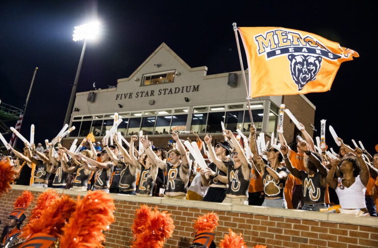 Fans in body paint cheer and wave sticks in front of Five Star Stadium, holding an orange Mercer Bears flag.
