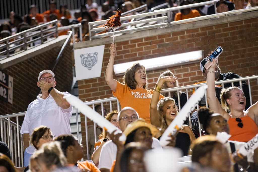 Mercer fans cheer in the stands at a sports event, some holding white noisemakers and pom-poms.