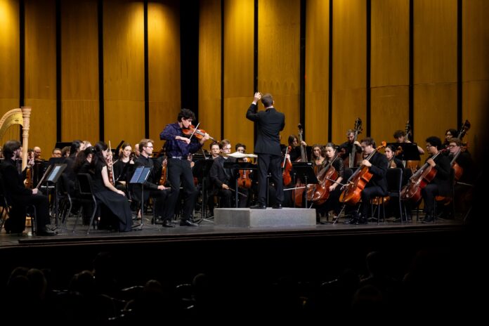 A conductor leads an orchestra on stage with a violin soloist performing at the front.