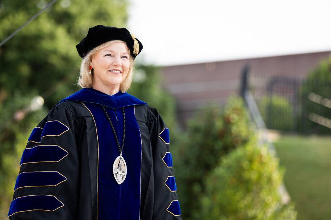 Woman in academic regalia and a cap stands outdoors, smiling, with greenery and a building in the background.