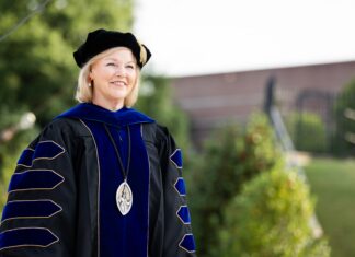 Penny L. Elkins, Ph.D., to be inaugurated as Mercer University’s 19th president, becoming its first woman president Woman in academic regalia and a cap stands outdoors, smiling, with greenery and a building in the background.