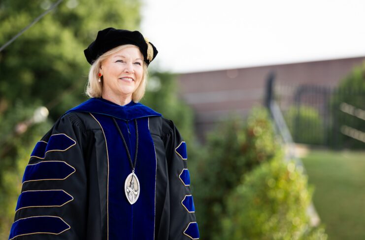 Woman in academic regalia and a cap stands outdoors, smiling, with greenery and a building in the background.