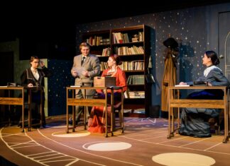 Mercer Theatre play honors female astronomy pioneers Four people in period costumes sit or stand at desks on a stage set resembling a vintage classroom with bookshelves.