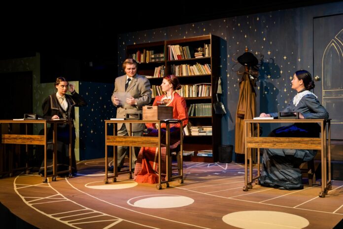 2026MercerTheatre-SilentSky_JessicaWhitley153_ Four people in period costumes sit or stand at desks on a stage set resembling a vintage classroom with bookshelves.
