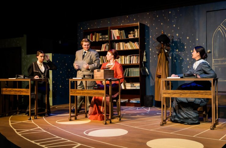 Four people in period costumes sit or stand at desks on a stage set resembling a vintage classroom with bookshelves.