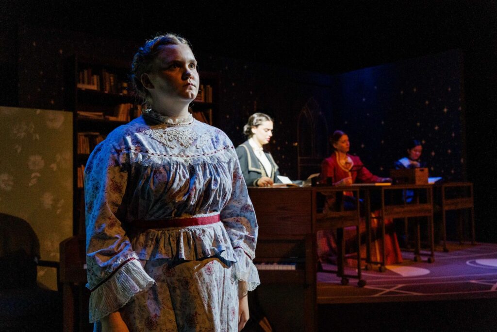A woman in a vintage dress stands in the foreground while three others work at desks in a dimly lit room.
