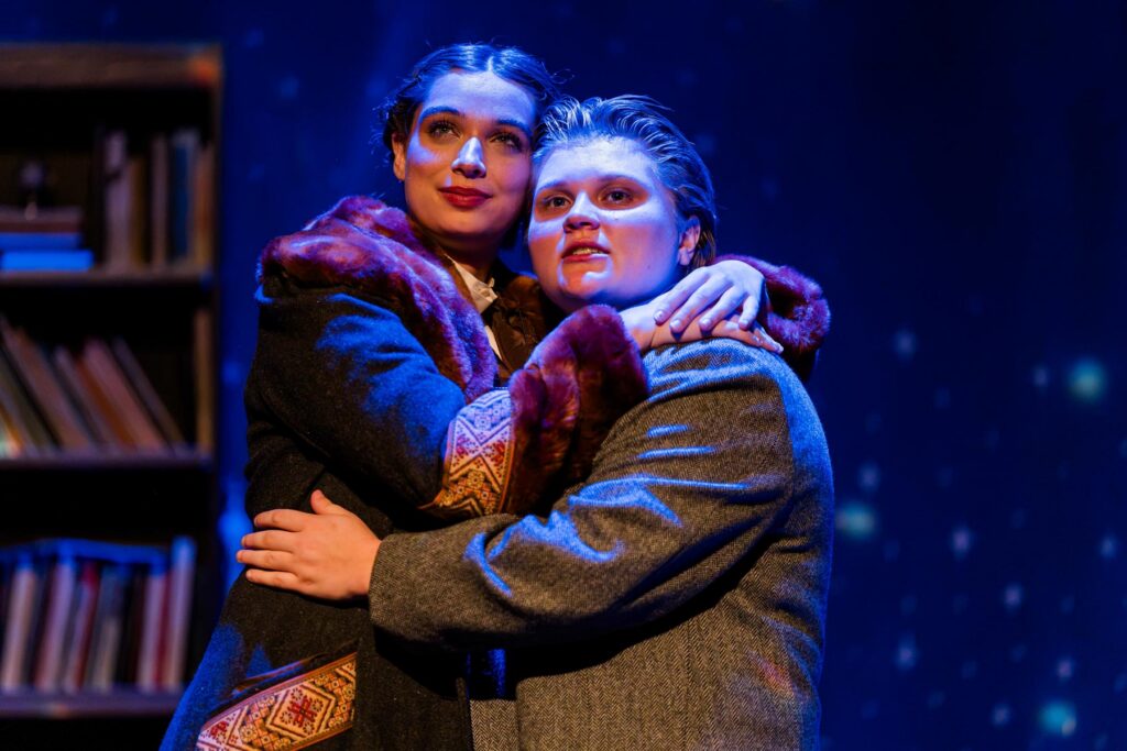 Two actors in vintage costumes embrace on stage with a starry blue backdrop and bookshelf on the left.