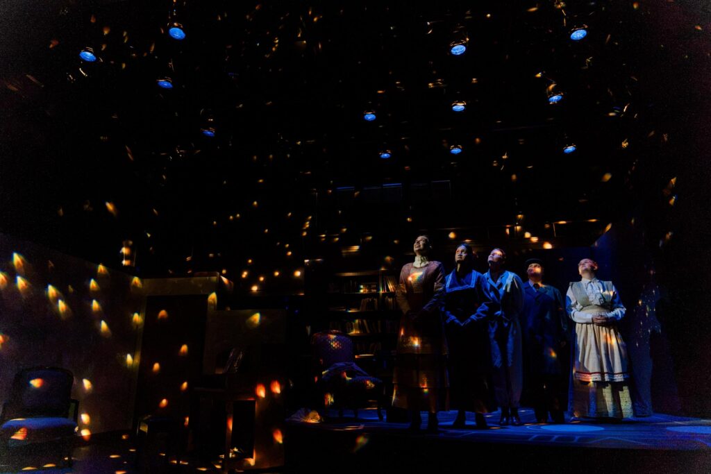 Four people stand on a dimly lit stage with scattered star-like lights illuminating the dark theater set.