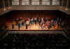 McDuffie Center for Strings presents 2026 Labor Day Festival A classical orchestra performs on stage in front of an audience in a concert hall with wooden paneling.