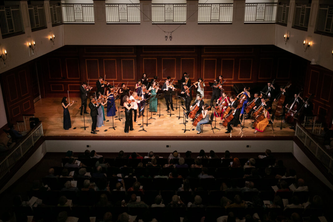 A classical orchestra performs on stage in front of an audience in a concert hall with wooden paneling.