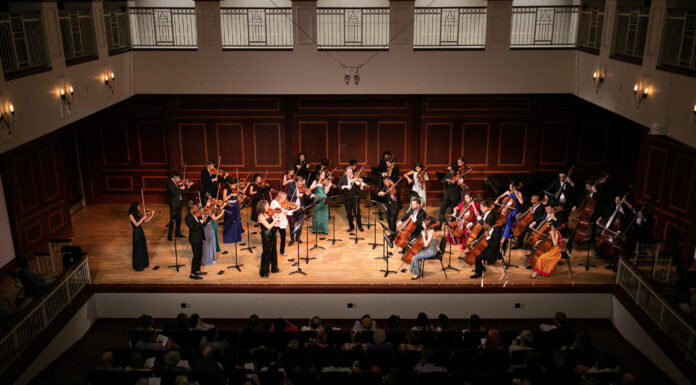 McDuffie Center for Strings presents 2026 Labor Day Festival A classical orchestra performs on stage in front of an audience in a concert hall with wooden paneling.
