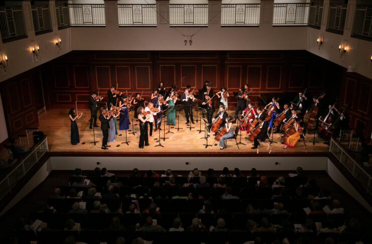 A classical orchestra performs on stage in front of an audience in a concert hall with wooden paneling.