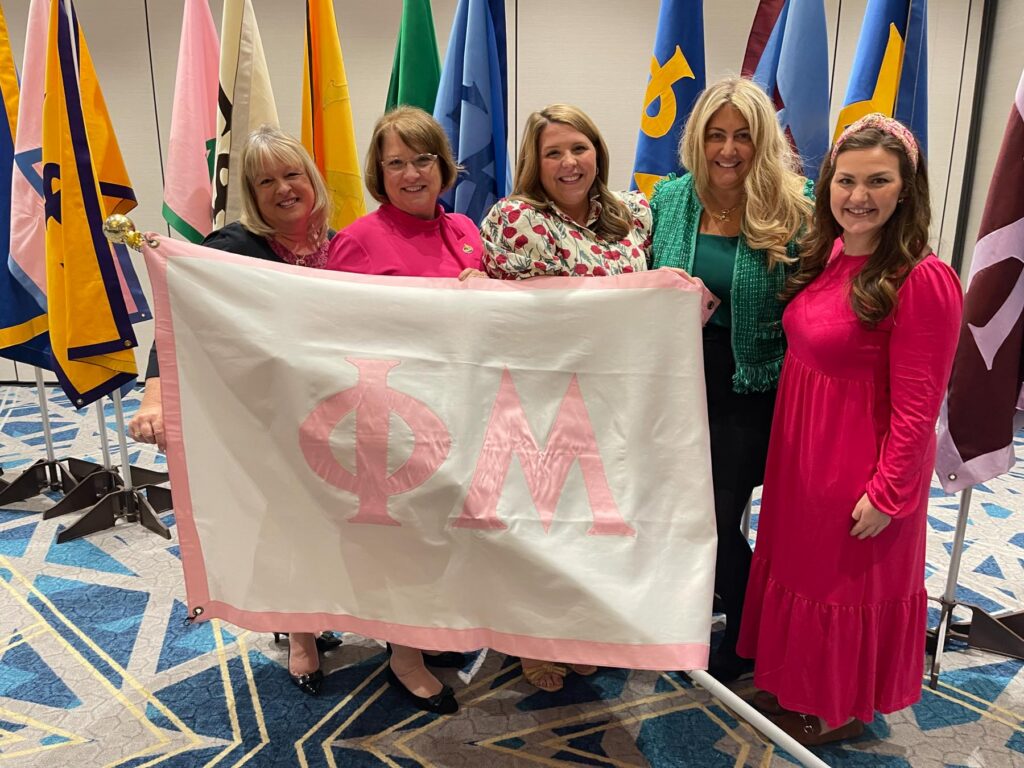 Five women stand together indoors holding a white and pink Phi Mu flag, with various flags displayed in the background.
