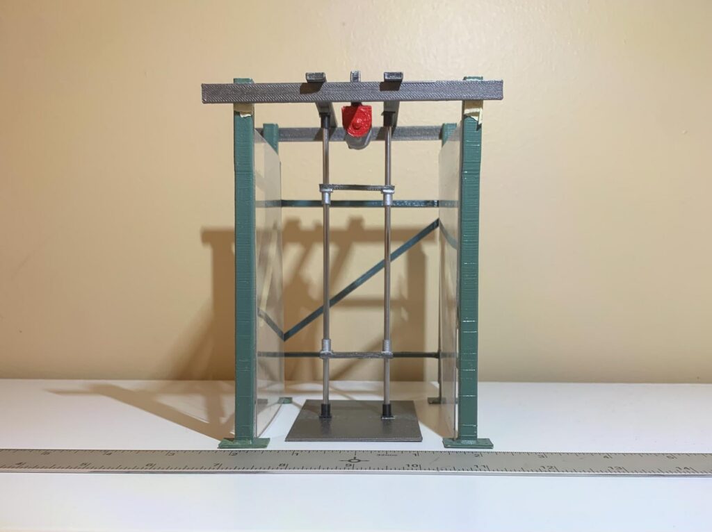 A small model structure with green pillars, a red component at the top, and a ruler in front on a white surface.