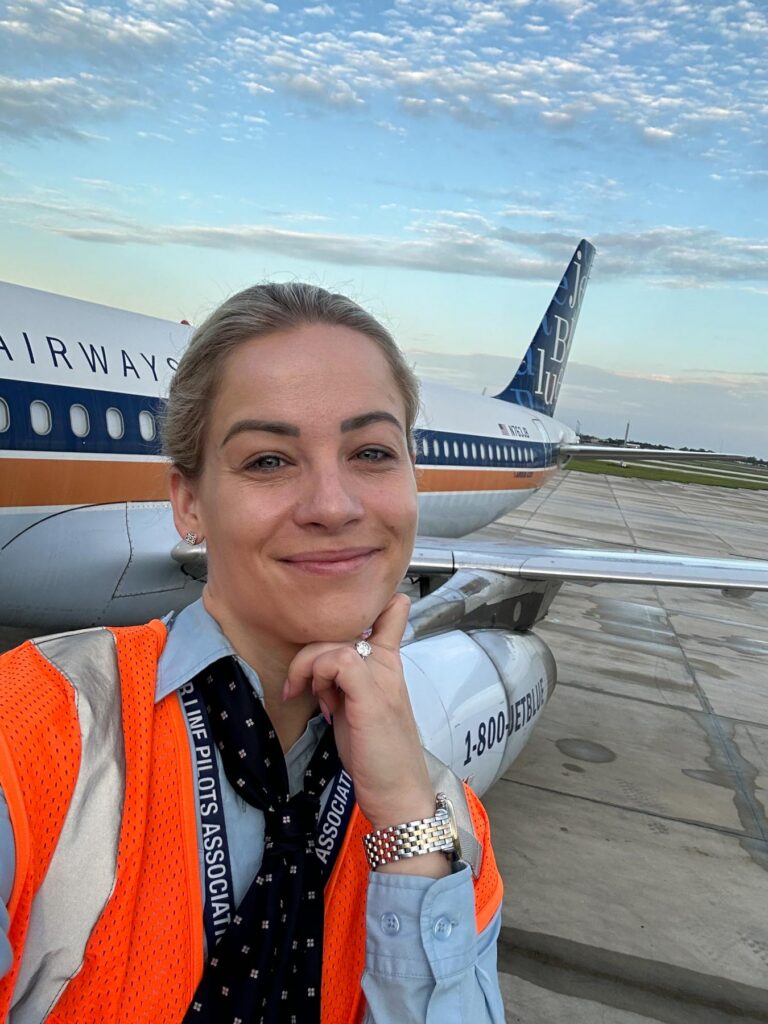 Person in safety vest poses in front of a parked JetBlue airplane on the tarmac under a partly cloudy sky.