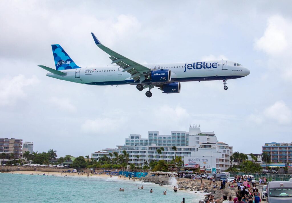 JetBlue airplane flying low over a crowded beach with hotels and palm trees in the background.