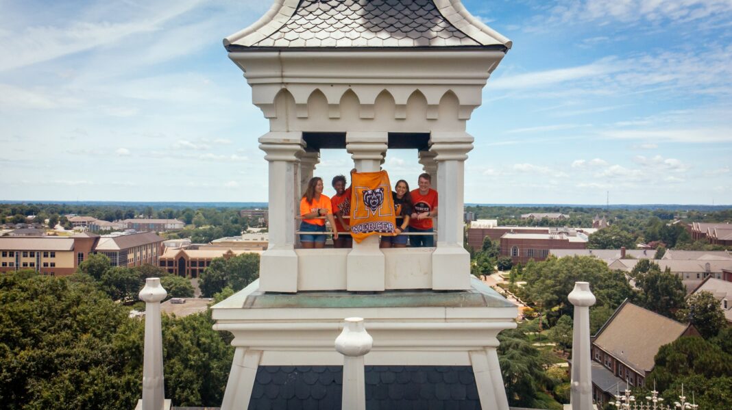 Four people stand in a tower holding a bright orange Mercer University Bears banner overlooking a college campus.