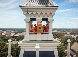 Mercer named a Best Value College for 2027 by The Princeton Review Four people stand in a tower holding a bright orange Mercer University Bears banner overlooking a college campus.
