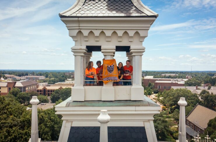 Four people stand in a tower holding a bright orange Mercer University Bears banner overlooking a college campus.