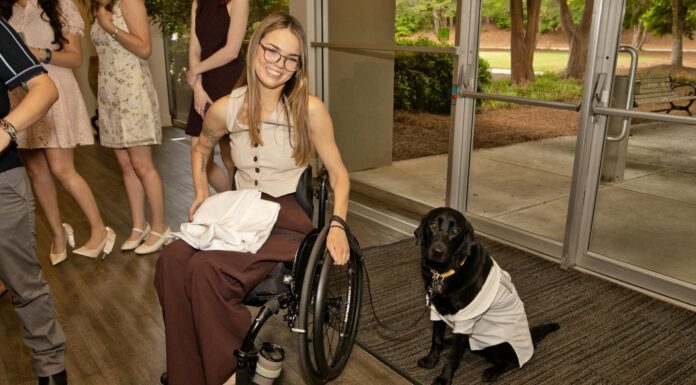 Doctoral student pursues passions in the classroom and the pool Woman in a wheelchair smiling beside a black dog wearing a white coat, indoors near glass doors.