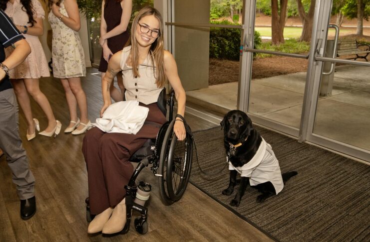 Woman in a wheelchair smiling beside a black dog wearing a white coat, indoors near glass doors.