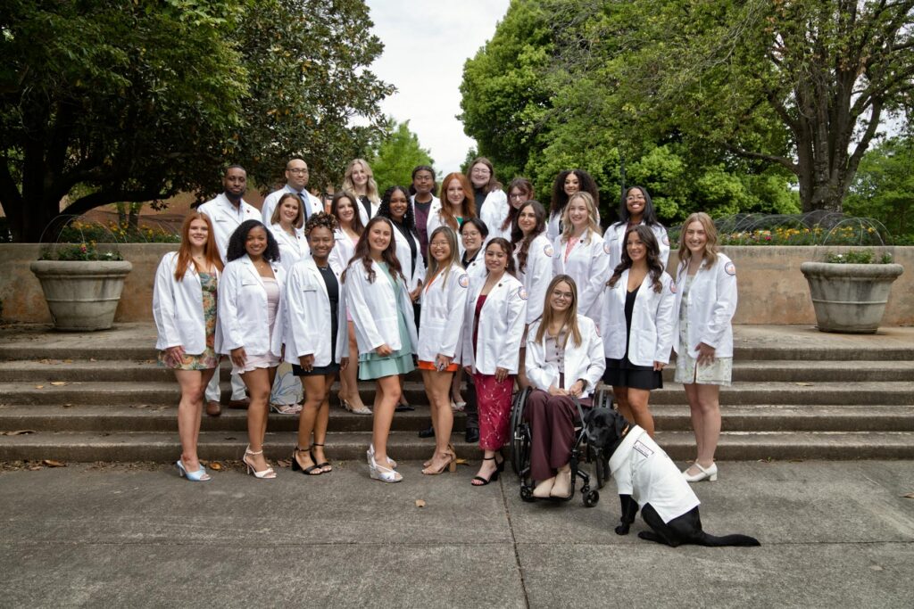 A group of people in white coats pose outdoors on steps; one person uses a wheelchair and a service dog is present.