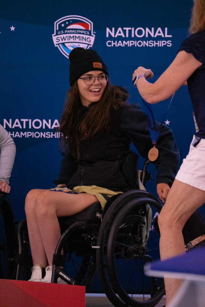 A young woman in a wheelchair smiles as she receives a medal at the U.S. Paralympics Swimming National Championships.