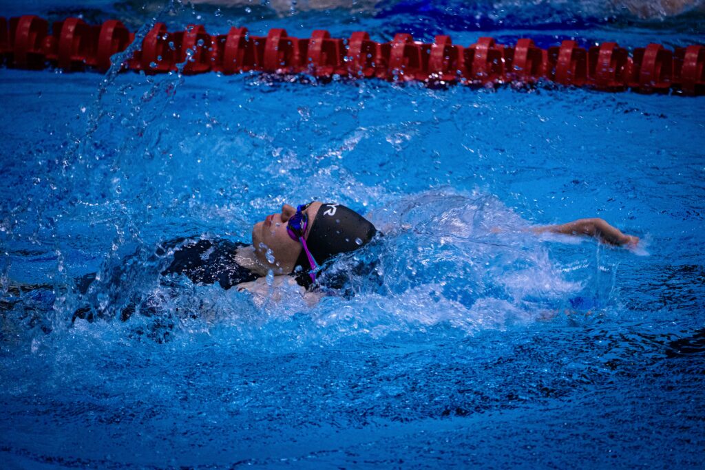 Swimmer in black suit and cap performing backstroke in a pool, water splashing around them near lane dividers.