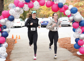 Mercer Law to host 9th annual Lauren Giddings 5K benefiting bar prep scholarships Two women with race bibs run under a balloon arch outdoors, passing orange cones on a paved path.