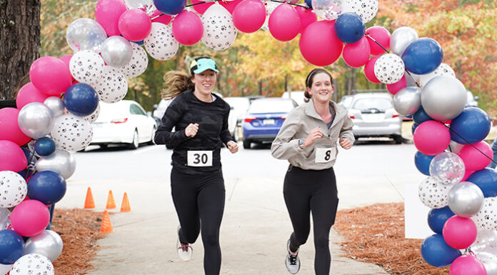 Mercer Law to host 9th annual Lauren Giddings 5K benefiting bar prep scholarships Two women with race bibs run under a balloon arch outdoors, passing orange cones on a paved path.