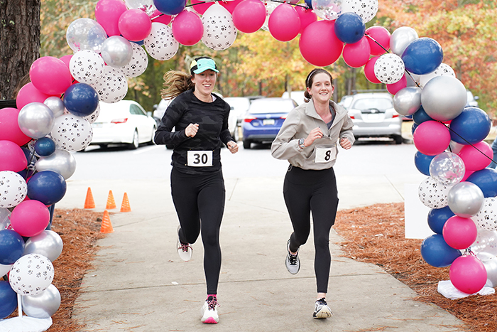 Two women with race bibs run under a balloon arch outdoors, passing orange cones on a paved path.