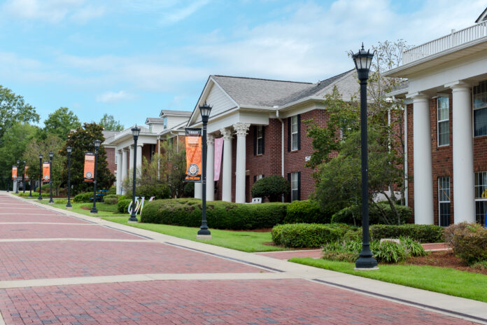 Brick walkways lead past Greek-style housing buildings with white columns and banners on a university campus.