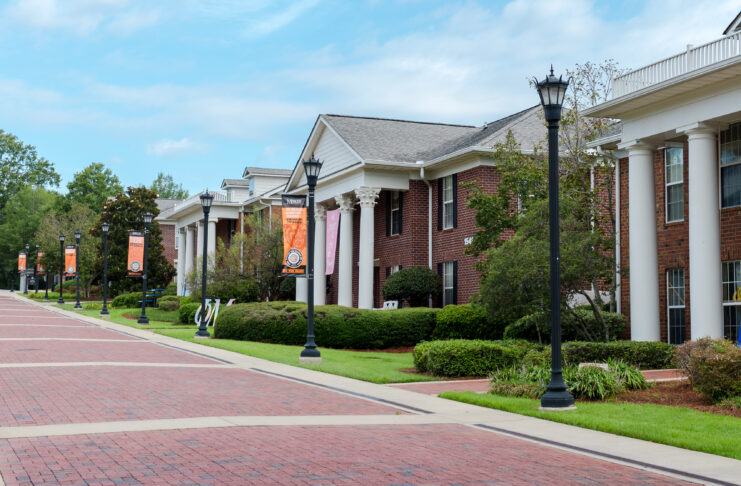 Brick walkways lead past Greek-style housing buildings with white columns and banners on a university campus.