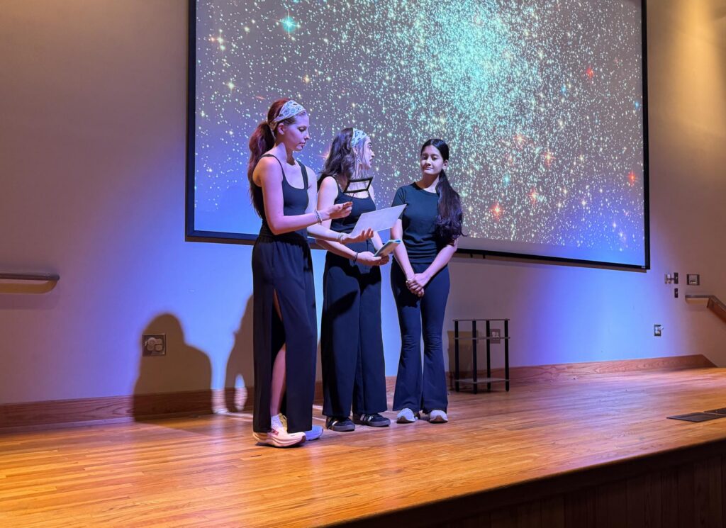 Three women stand on a stage, one holding papers, in front of a starry projection background.