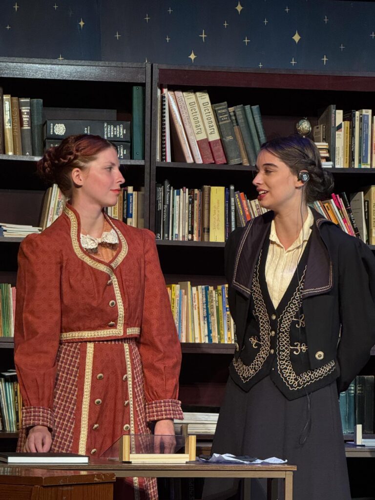 Two women in period costumes stand in front of bookshelves, appearing to converse on a stage set resembling a library.