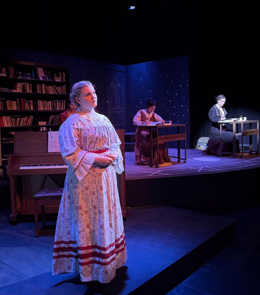 Three women in period costumes perform on a dimly lit stage set resembling a library and study.