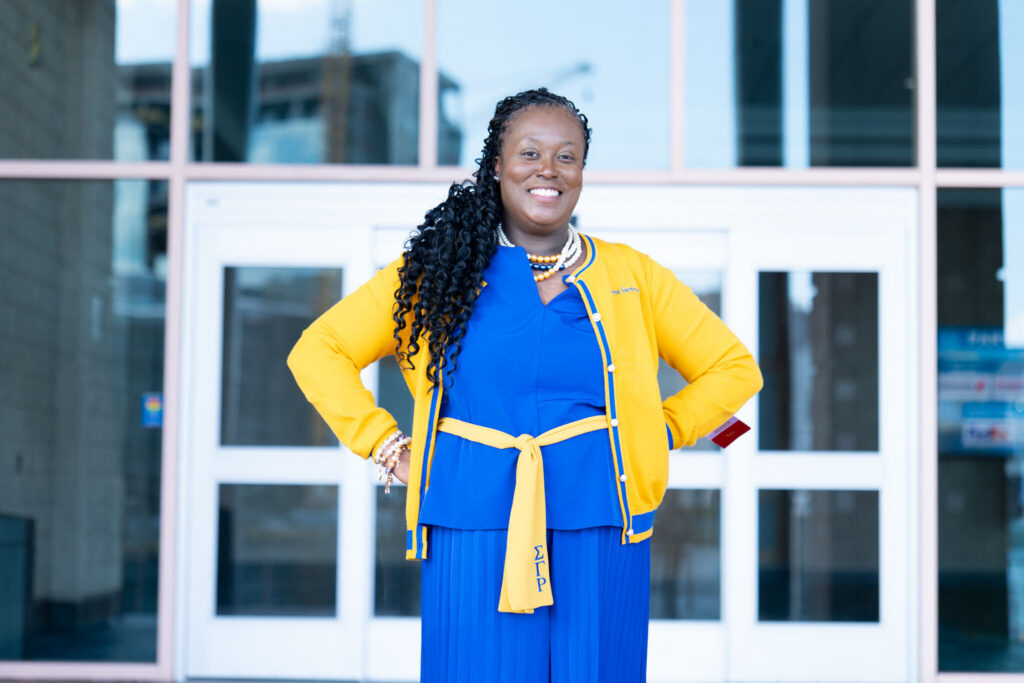 Woman standing in front of glass doors, wearing a blue outfit with a yellow jacket, smiling with hands on hips.