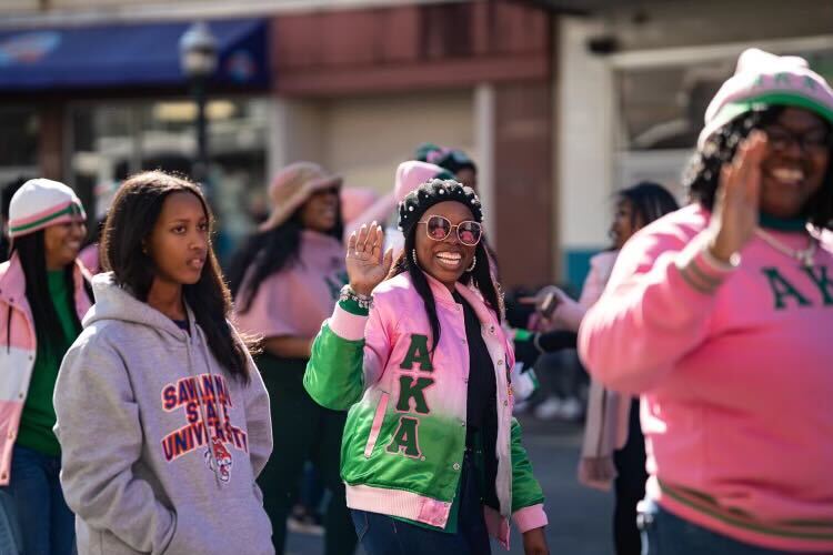 Group of women, some wearing pink and green jackets, walk outdoors; one woman waves and smiles at the camera.