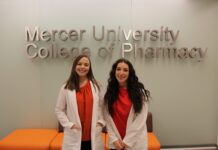 Pharmacy students dive into research through summer program Two women in white coats stand and smile in front of a Mercer University College of Pharmacy sign.