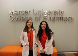 Pharmacy students dive into research through summer program Two women in white coats stand and smile in front of a Mercer University College of Pharmacy sign.