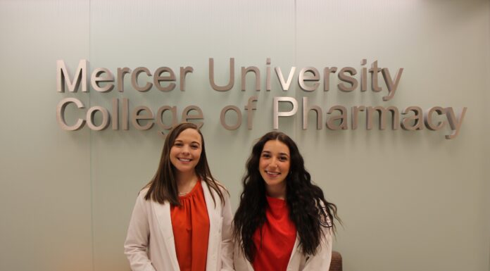 Pharmacy students dive into research through summer program Two women in white coats stand and smile in front of a Mercer University College of Pharmacy sign.