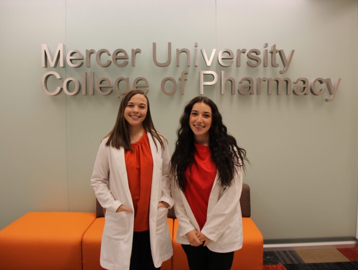 Two women in white coats stand and smile in front of a Mercer University College of Pharmacy sign.