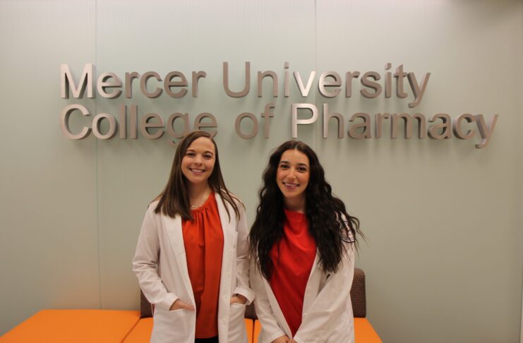 Two women in white coats stand and smile in front of a Mercer University College of Pharmacy sign.