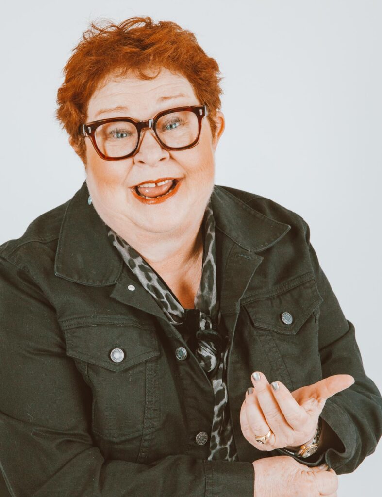 Smiling person with short red hair, glasses, and a dark jacket gestures with one hand against a plain light background.