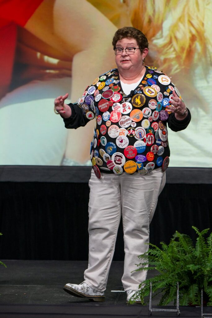 Person standing on stage wearing a black jacket covered in assorted colorful pins and buttons, with plants in the foreground.