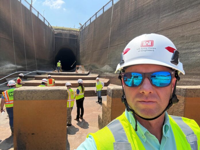 Rich 2-2 A group of workers in safety vests and helmets stand near a large tunnel under construction; one man is in the foreground.